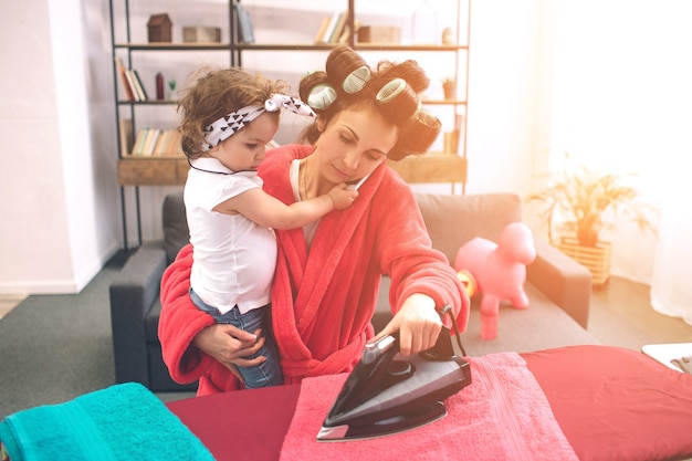 Mother and baby together engaged in housework Ironing clothes . Housewife and kid doing homework. Woman with little child in the living room. Homemaker doing many tasks while looks after her infant.