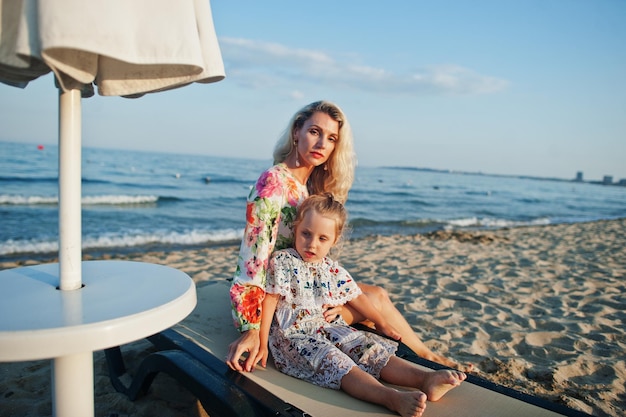 Mother and beautiful daughter having fun on the beach sitting on sunbed Portrait of happy woman with cute little girl on vacation