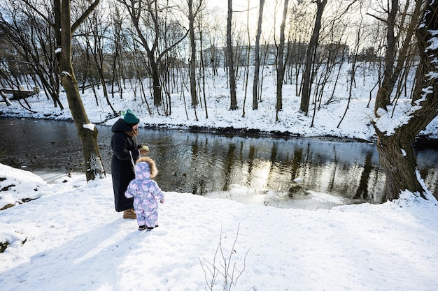 Mother and child walking on a sunny frosty winter day in the park near river with ducks and birds