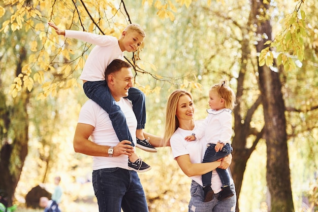 Mother and dad holds kids at shoulders and in hands. Cheerful young family have a walk in an autumn park together.