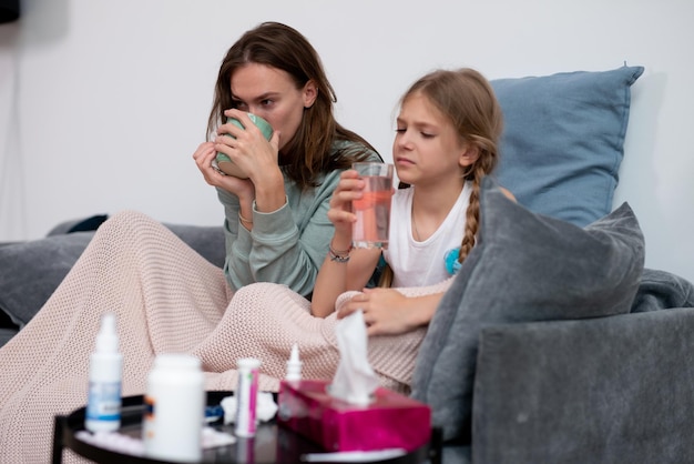 Mother and daughter are sick together sitting on the sofa and drinking medicine