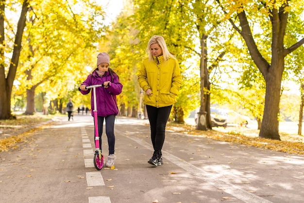 Mother and daughter are walking in the autumn in the park. Little girl is riding a scooter