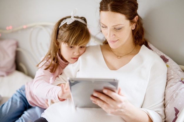 Mother and daughter looking at tablet, sitting on bed.