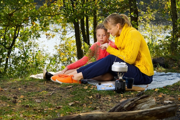 Mother and daughter play on a blanket in the autumn forest near the river or lake. Young mother playing with her daughter in autumn park on sunny day