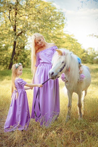 Mother and daughter in similar lavender dresses are petting a unicorn horse. Summer meadow