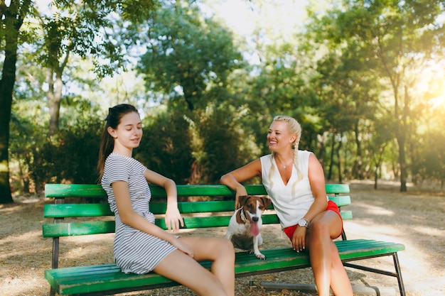 Mother and daughter sitting on the bench with small funny dog. Little Jack Russel Terrier pet playing outdoors in park. Dog and women. Family resting on open air.