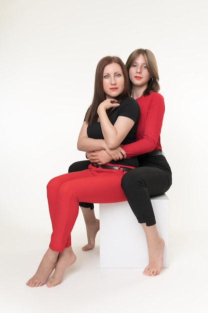 Mother and daughter sitting on white cube box on white background Daughter sits behind hugging mom