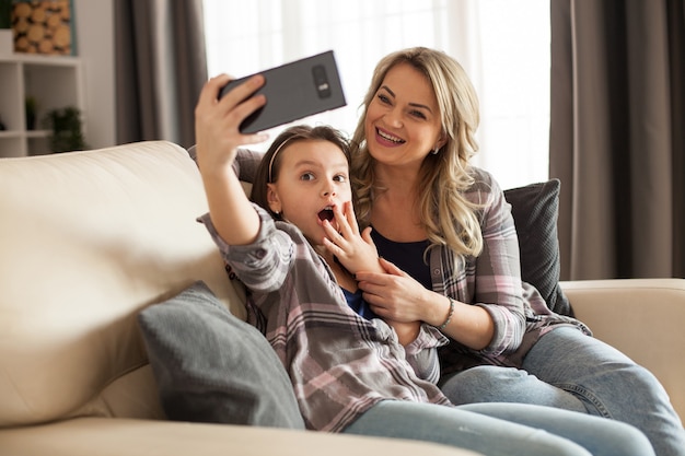 Mother and daughter in their apartment sitting on the couch smiling and taking a selfie.