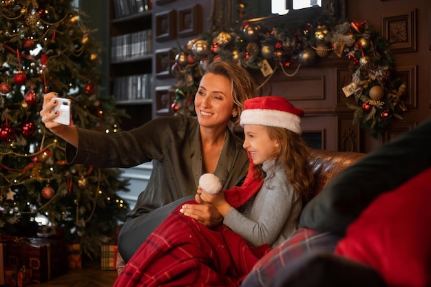 Mother and daughter wearing Santa hat taking selfie in Christmas decorated living room