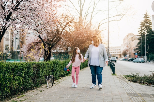 Mother and daughter with protective masks on their faces enjoying in walk in park with their dog.