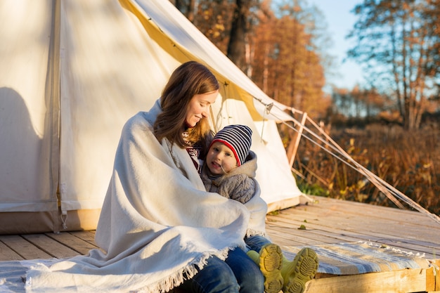 Mother embracing her kid with a blanket while sitting near camping tent