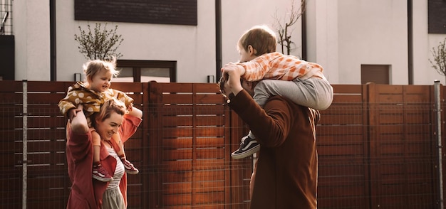 Mother father carrying children on piggyback near house on sunset