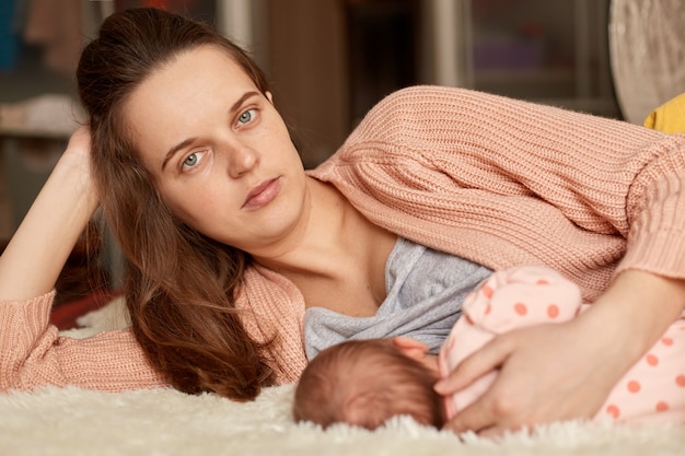 Mother feeding breast her infant, european woman posing with new born baby while lying in bed on fluffy blanket, female with dark hair looks at camera.