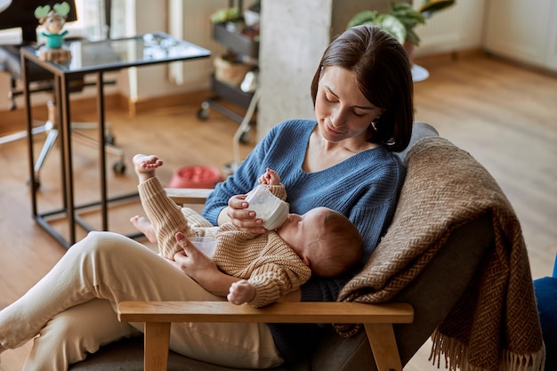 Mother feeding her newborn baby from bottle with milk. Young european woman and her infant kid sitting in wooden armchair. Interior of studio apartment. Concept of motherhood and child care