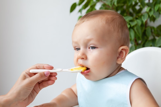 Mother feeds funny happy blond blue eyed baby in blue bib from a spoon