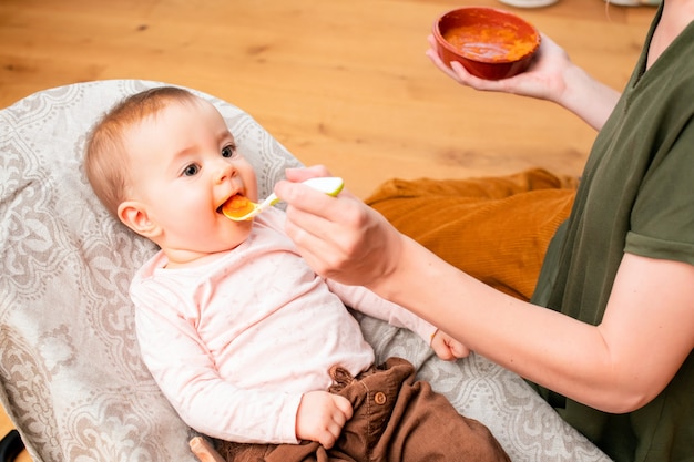 Mother feeds smiling baby vegetable puree from a plastic spoon at home.