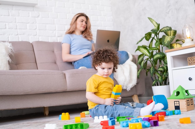 Mother freelancer use laptop sits on couch kid daughter plays with toys on floor working from home