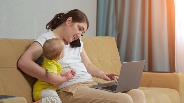 Mother freelancer works on laptop talks on phone holding kid