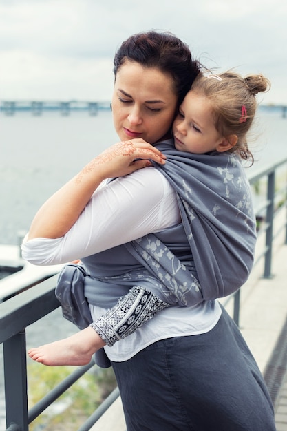 Mother and girl wrapped in grey sling back at city river background, baby wearing