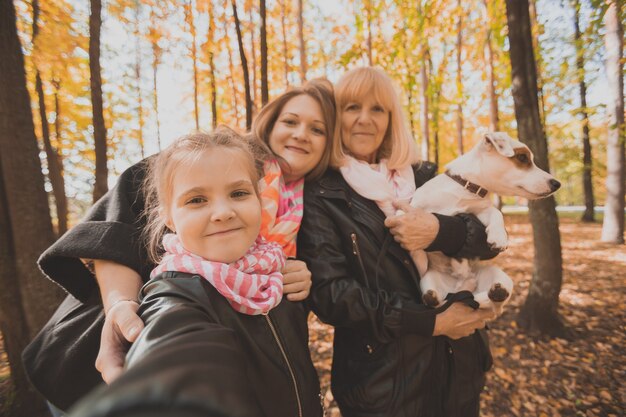 Mother, grandmother and little granddaughter with jack russell terrier dog taking selfie by smartphone outdoors in autumn nature. Family, pets and generation concept