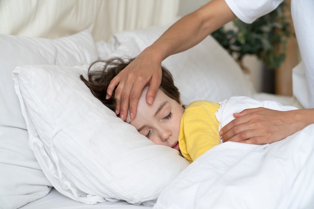 Mother hand touch boy forehead check temperature of sick son sleep in bed with fever from influenza