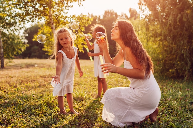 Mother helps daughters to blow bubbles in summer park