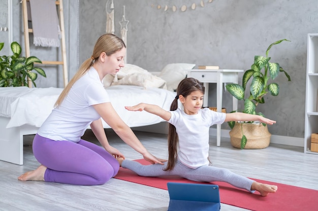 mother helps her daughter doing stretching exercising online with tablet at home