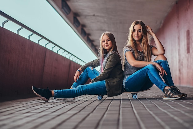 Mother and her daughter schoolgirl with blonde hair dressed in trendy clothes sitting together on a skateboard at bridge footway.