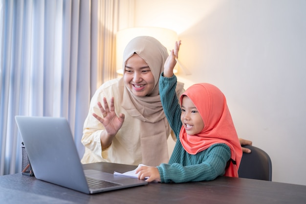 Mother and her daughter wave her hand to laptop computer during class meeting with school from home