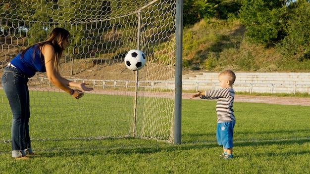 Mother and her small son standing in front of goalposts on a green grass sportsfield throwing a soccer ball to each other