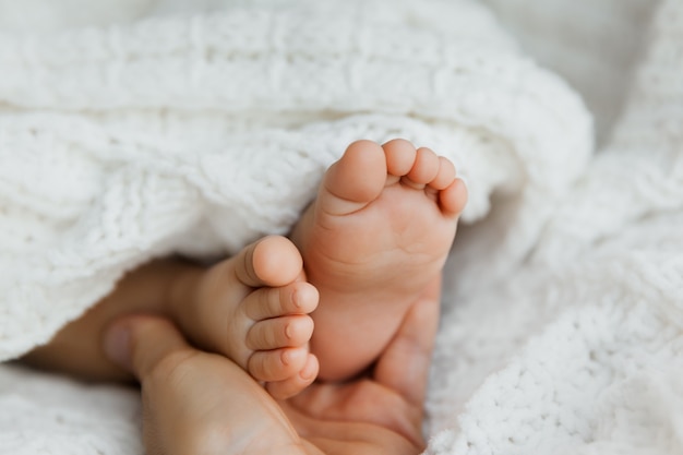 Mother holding feet of newborn daughter, closeup of infant toes on white blanket