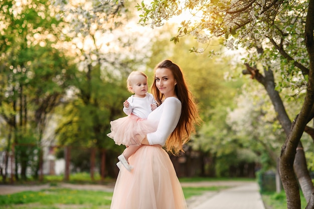 Mother holds her little daughter in her arms among blooming trees. Mom and her little baby weared pink family look dress.