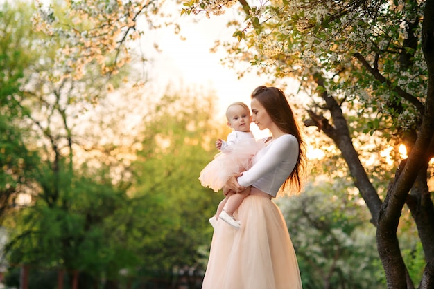 Mother holds her little daughter in her arms among blooming trees. Mom and her little baby weared pink family look dress.