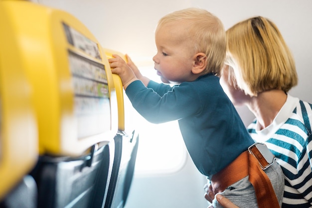 Mother and infant baby boy child flying on comercial flight traveling by plane sitting together at window seat Flying with kids