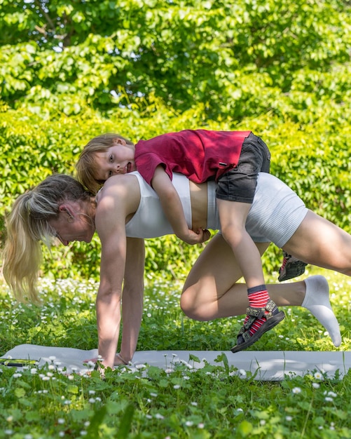 mother is engaged in sports exercises with her son in the fresh air The child sits on the back of h