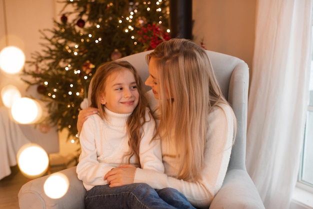 Mother and kid girl daughter celebrating christmas holidays sitting in chair over xmas tree in room Winter greeting season Celebration Motherhood