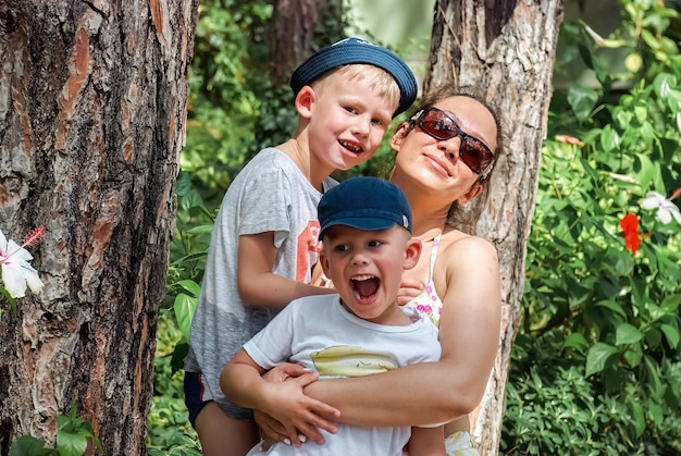 Mother and little boys stand leaning on pine trunks in hotel park