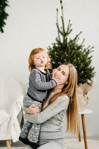 Mother and little child having fun and playing together at home Portrait loving family close up Cheerful mom hugging cute baby daughter girl near Christmas tree Merry Christmas and Happy Holidays