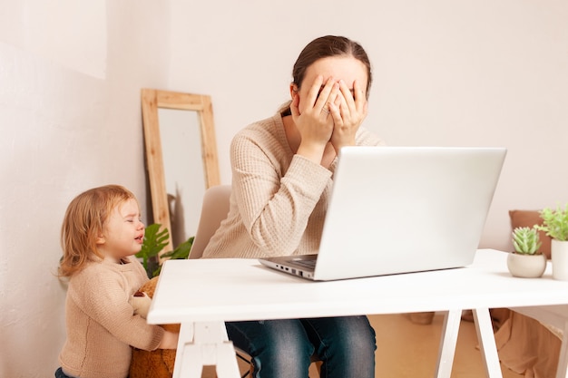 A mother on maternity leave sits at a laptop and works a child yelling hysterically cries distracts