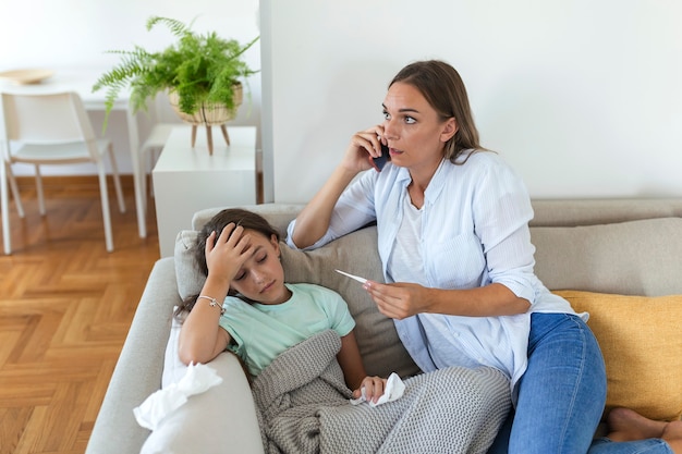 Mother measuring temperature of her ill kid. Sick child with high fever laying in bed and mother holding thermometer. Mother with cell phone calling to doctor