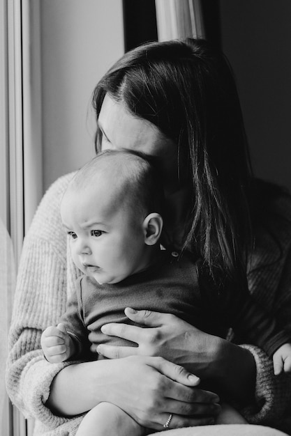 Mother playing with baby son at home near the window. Happy infant and mom.