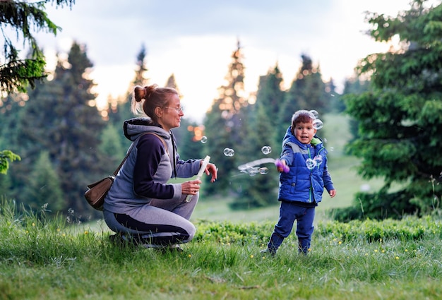 Mother plays with son on a lawn and teaches him to blow soap bubbles against the backdrop of forest