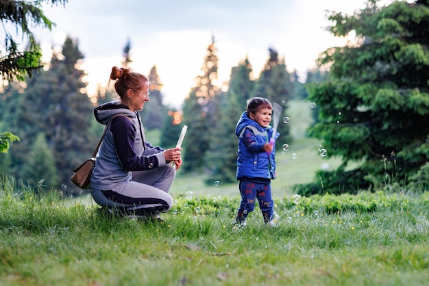 Mother plays with son on a lawn and teaches him to blow soap bubbles against the backdrop of forest