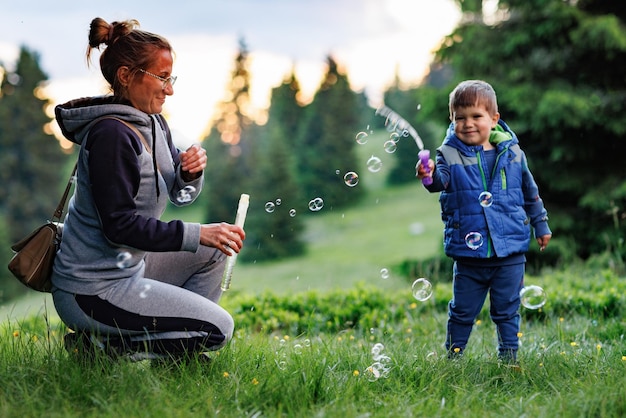 Mother plays with son on a lawn and teaches him to blow soap bubbles against the backdrop of forest