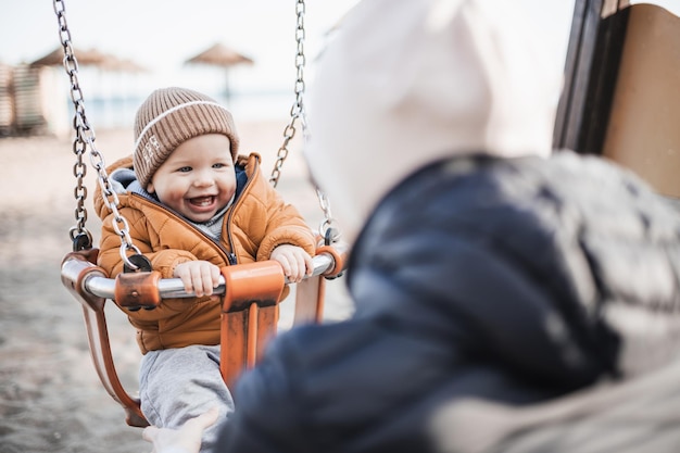 Mother pushing her cheerful infant baby boy child on a swing on sandy beach playground outdoors on nice sunny cold winter day in Malaga Spain