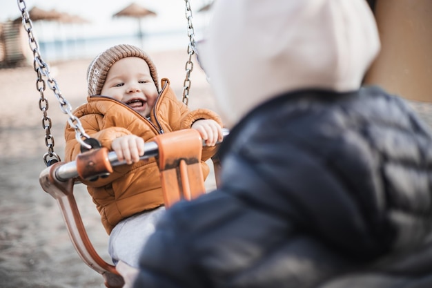 Mother pushing her cheerful infant baby boy child on a swing on sandy beach playground outdoors on nice sunny cold winter day in Malaga Spain