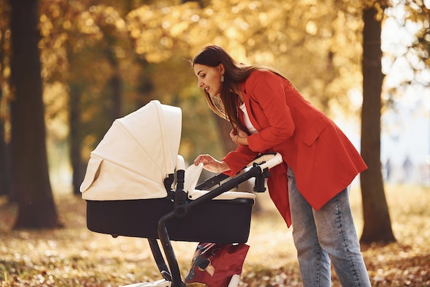 Mother in red coat have a walk with her kid in the pram in the park with beautiful trees at autumn time.