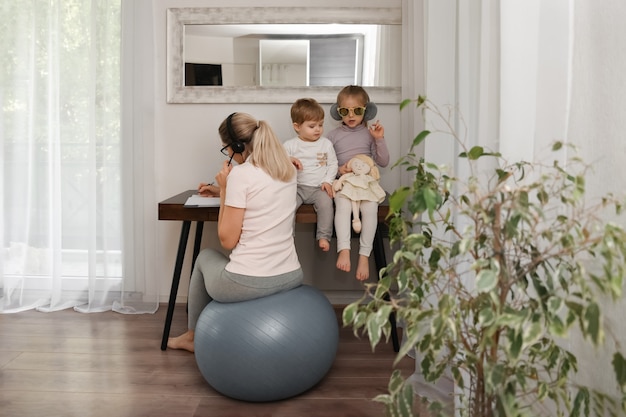 Mother sits on a fitness ball in headphones and works at home with her children during quarantine Covid-19