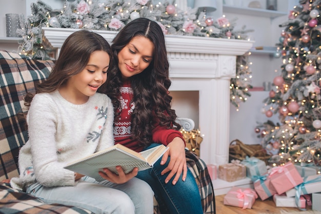 Mother sittng together with daughter and look at book. They read it. Girl holds it with both hands. They are in decorated room. There are Christmas tree and fireplace behind them.