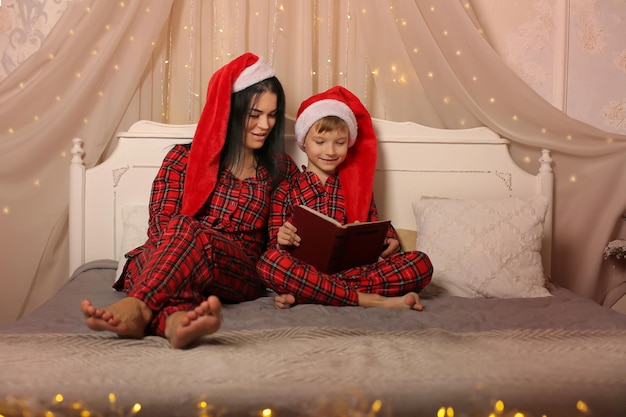 mother and son in christmas pajamas santa Claus hats reading a book sitting on the bed